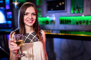 Smiling woman holding white wine glass at bar counter with neon green backlight and bottle shelves
