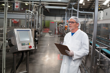 Senior man wearing lab coat inspecting conveyor at factory with clipboard at panel, copy space