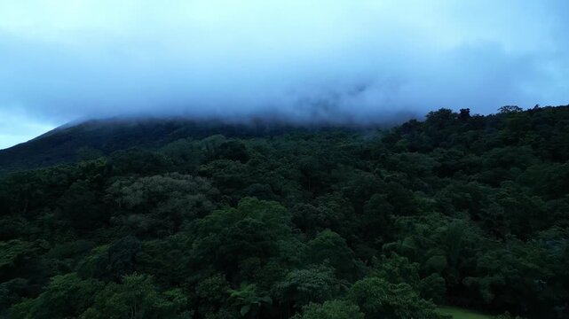 A sky-high shot glides over dense tropical forest towards the mist-covered Mount Banahaw, capturing rich greenery under a moody, clouded sky in Quezon Province Philippines