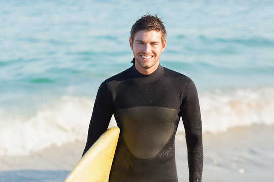 Male surfer standing at edge of sandy beach wearing black wetsuit and holding yellow surfboard