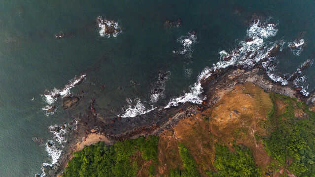 Aerial drone top view of Vagator Beach in Goa, India, with waves crashing on the shore, scattered rocks in the Arabian Sea, and lush green trees framing the scenic coastline.