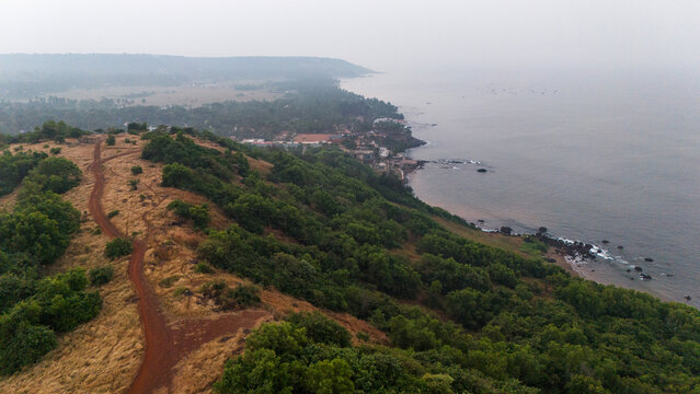 Aerial shot of hilltop at Vagator Beach, Goa, during winter with yellow grass, green trees, brown mud paths, and the Arabian Sea with rocks and ocean waves.