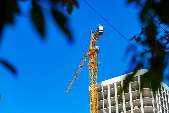 Against a clear blue sky, a tower crane works on a multi-story building, its yellow structure standing out against the concrete columns and ceilings. - Powered by Adobe