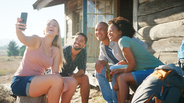 Group Of Friends Posing For Selfie On Mobile Phone Sitting On Steps Of Cabin In Countryside