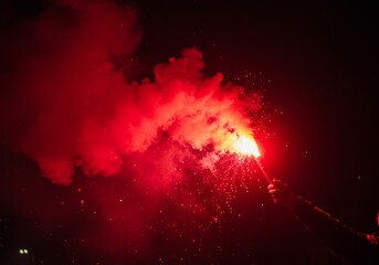 Hand holding a burning red flare with smoke and sparks.