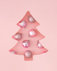 Winter composition with Xmas baubles visible through a Christmas tree shaped gap on pastel pink background. Minimal Christmas and New Year concept. Creative festive holiday layout. Flat lay, top view.