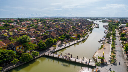 An aerial view of Hoi An's old town and river, showing the rows of historic houses and a long line of tourist boats, with a scenic backdrop of mountains and the modern city.