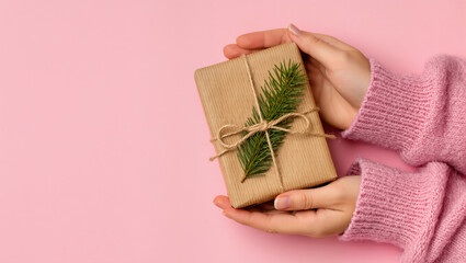 Christmas gift in eco wrapping with fir twig and twine held in female hands in pink sweater on pastel pink background