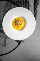 Creamy yellow risotto served in a white bowl, garnished with herbs and vegetables. Elegant minimalist presentation on a glass table with wicker chair in background