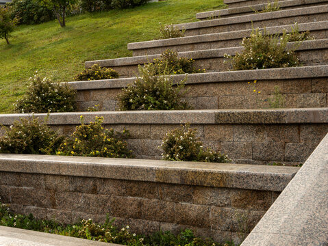 Modern landscape architecture featuring terraced retaining walls of natural stone. The wide, tiered steps are integrated with green shrubs and plants, creating a harmonious public garden space.