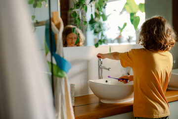 A child stands at a stylish bathroom sink, brushing their teeth. Morning light fills the space, creating a fresh and inviting atmosphere with greenery in view. © m-art