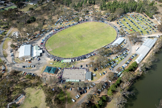 Country football match in progress with crowd lining the oval boundary
