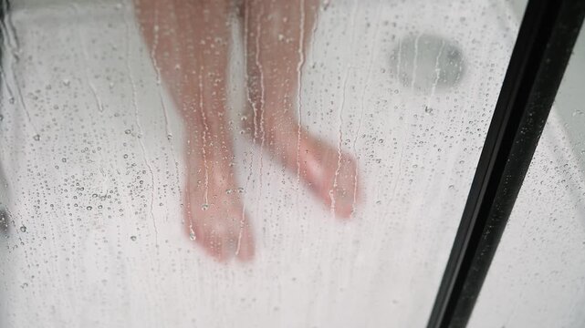 Wet, bare feet standing behind glass shower door, water droplets covering transparent surface during personal hygiene routine. Selective focus
