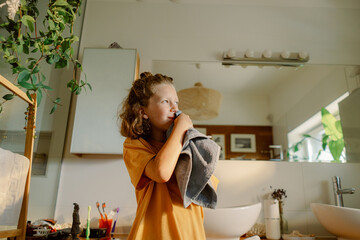 A child stands in a warm, inviting bathroom, holding a towel close while getting ready for bath time. The space features plants and a stylish design, perfect for relaxation.