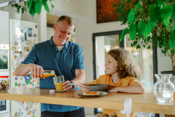 A father pours juice for his child at a kitchen counter. The cheerful atmosphere is filled with warm light as they enjoy their morning routine, creating lasting memories together.