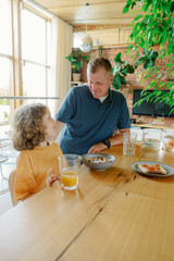 A parent and child share a joyful breakfast at a modern dining table. They engage in conversation while surrounded by warm natural light and green plants, creating a cozy atmosphere.