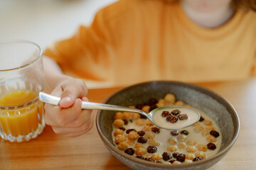 A child holds a spoon with cereal and milk while sitting at a wooden table. A glass of orange juice sits nearby, with sunlight streaming through the window creating a warm atmosphere.