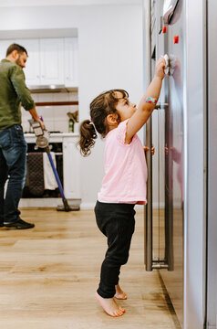 Toddler girl cleaning the fridge door while father vacuums in the background