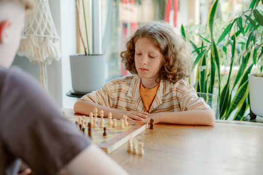 A boy focused on a chess match occupies a cozy indoor space, surrounded by plants. He is playing with his father or brother, engaged in a moment of strategic thinking.