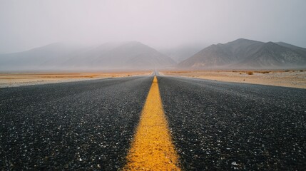Empty asphalt road with yellow line leading towards misty mountains under a cloudy sky