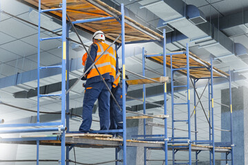 Ventilation and air conditioning installation. Workers in helmets and reflective vests stand on scaffolding inside a building, assembling and maintaining duct systems for climate control.