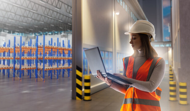 Logistics management. Female worker in safety vest and helmet uses a laptop in a warehouse with empty racks, controlling supply chain operations and warehouse processes. - Powered by Adobe