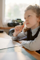 A young boy sitting at a table is deep in thought as he considers his schoolwork. A pencil is held thoughtfully to his lips, showcasing his focus and imagination.