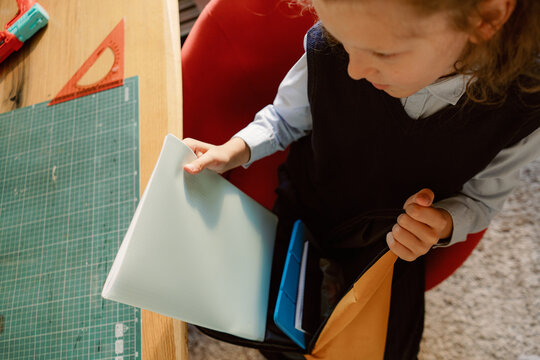 A young boy sits at a stylish table examining his school supplies with focus. His backpack is open nearby, showing preparation for a busy school day. The setting reflects a cozy study area.