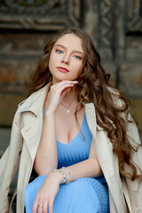 Fototapeta premium Close-up portrait of a young stylish woman with long wavy hair, wearing a blue dress and beige trench coat, looking confident outdoors. Elegant fashion and lifestyle photography