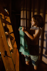 In a dimly lit room, a young boy examines a piece of clothing, showcasing his morning routine. Sunlight casts soft shadows, creating a warm atmosphere as he prepares for the day ahead.
