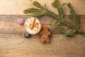 Christmas holiday traditional eggnog topped with spices and a cinnamon stick, on wooden table with gingerbread cookies, bakers' twine and jingle bell.