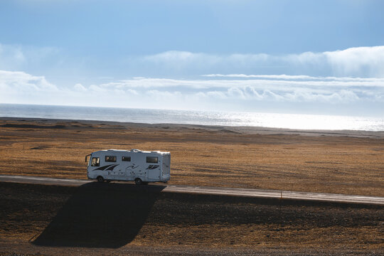 On the way to Fjallsarlon. Serene view of lonely trailer going on the narrow road in wasteland along empty seashore as sun shining brightly. Calm Arctic Ocean and clear blue sky seen in background.