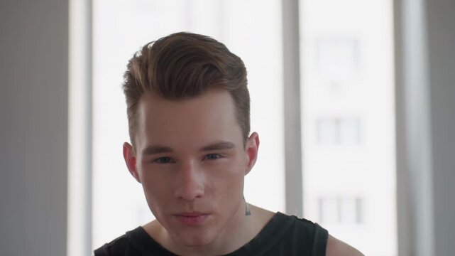 handsome tattooed man in sleeveless signet sits before bright window mirror, holds black makeup sponge, delicately applies powder to cheeks with poised expression