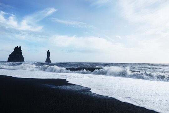 Black sand beach of Vik. Gorgeous view of Arctic Ocean waves crashing on black volcanic sand shore. Black rock formations sticking out of ocean in distance and birds soaring high in clear blue sky.