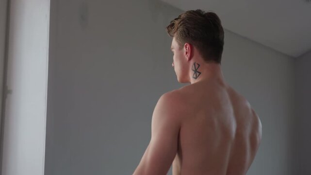 shirtless man with neck tattoo performing resistance rope exercise near large window in minimalist indoor setting, showcasing upper body strength and muscular back during intense training routine