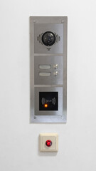 Metal Intercom Panel on a White Wall Featuring a Camera, Two Buttons, and a Key Card Reader with an Illuminated Indicator Light Below a Red Emergency Button