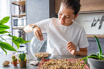 Young woman pouring stones into a glass container while preparing a terrarium. Concept of mindful crafting, eco-friendly lifestyle and home plant design.