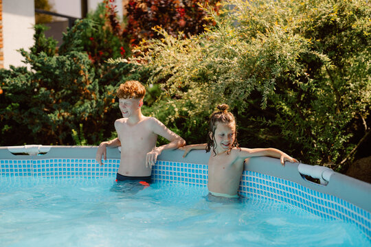 A young boy and his brother play in their backyard pool during a warm, sunny day. They relax against the pool edge, laughing and enjoying their summer afternoon together.
