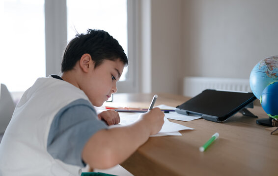 Schoolboy doing homework and focused writing on paper at desk with tablet and globe, symbolizing education, representing home learning, studying, e-learning, and modern digital classroom at home