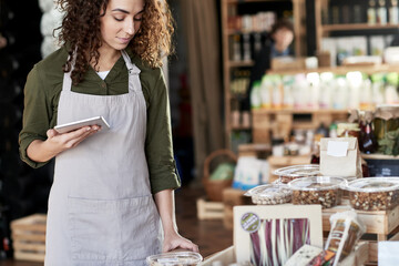 Portrait of young pretty curly shop assistant in apron working in organic food store. She standing at the counter piled with natural products, holding tablet, looking down and smiling slightly.