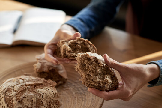 Extreme close-up of female hands in denim shirt holding big pieces of homemade freshly-baked whole wheat crusty bread. Rest of loaf lying on a wooden table next to notepad with pen.