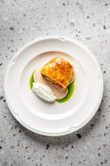 Gourmet plated dish with puff pastry, creamy quenelle, brown sauce, and green oil on white ceramic plate. Minimalist fine dining presentation on textured gray background