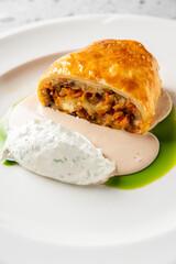 Gourmet plated dish with puff pastry, creamy quenelle, brown sauce, and green oil on white ceramic plate. Minimalist fine dining presentation on textured gray background
