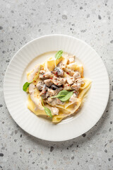 Pappardelle pasta with creamy chicken and mushroom sauce, garnished with basil and chili flakes. Served on a white plate over a light stone background. Top view, close-up