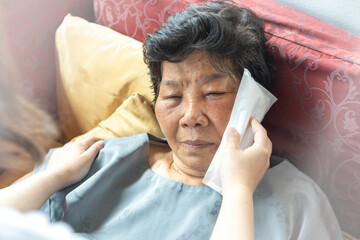 Elderly woman lying on sofa with caregiver applying an ice pack to her face, symbolizing family care, home healthcare, pain relief, swelling management, headache, and tooth problem treatment.