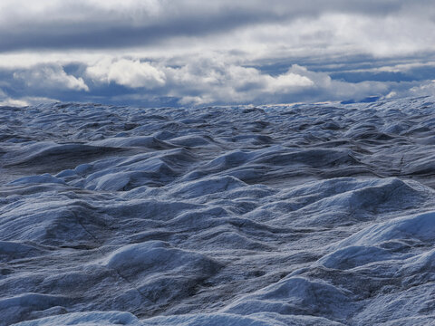 Wavy blue ice surface of glacier with cloudy sky, showing natural textures and frozen terrain.