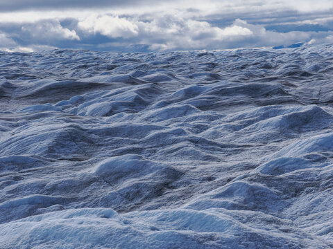 Wavy blue ice surface of glacier with cloudy sky, showing natural textures and frozen terrain.