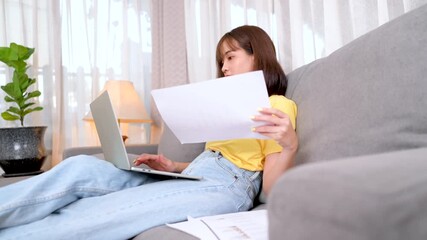 A young woman working from home on a laptop while reviewing printed documents, symbolizing remote work, freelance career, financial analysis, and modern digital lifestyle. - Powered by Adobe