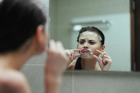 Discontented Caucasian woman applying facial mask in bathroom mirror, expressing frustration while caring for her skin. Beauty and self-care concept.