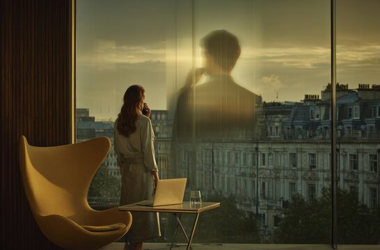 Rear view of a woman in robe holding mug, gazing out from a city-view suite. Laptop and drink on table with yellow chair. Sunlight